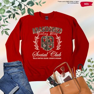 May include: A red sweatshirt with the text "ABANDONED Hunting Wives Social Club" in a camouflage design. The sweatshirt features a crest with hunting-themed graphics and the phrase "HELLO HUNTING SEASON, GOODBYE HUSBAND."