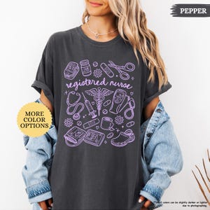 May include: Dark gray t-shirt with a lavender graphic of medical tools and the words "registered nurse." The design includes a stethoscope, syringe, and other medical equipment. A button on the left says "MORE COLOR OPTIONS."