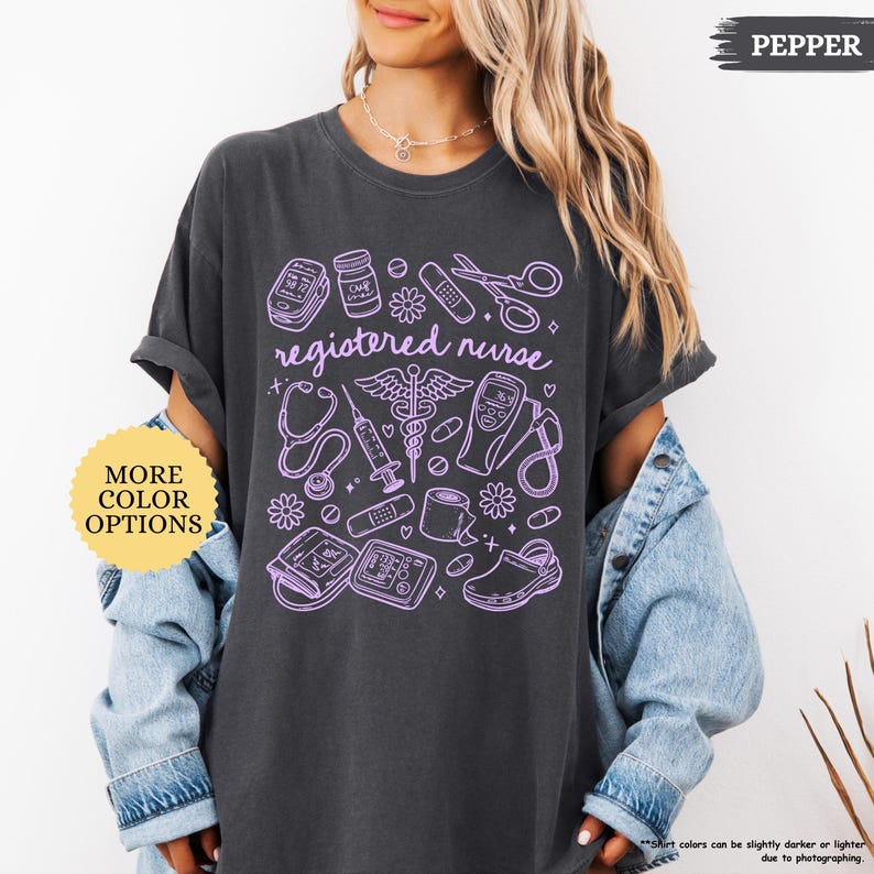 May include: Dark gray t-shirt with a lavender graphic of medical tools and the words "registered nurse." The design includes a stethoscope, syringe, and other medical equipment. A button on the left says "MORE COLOR OPTIONS."
