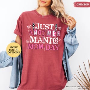 May include: Crimson-colored t-shirt with the text "JUST ANOTHER MANIC MOM-DAY" in a pink and white design. The shirt has short sleeves and a relaxed fit. A "MORE COLOR OPTIONS" button is visible.