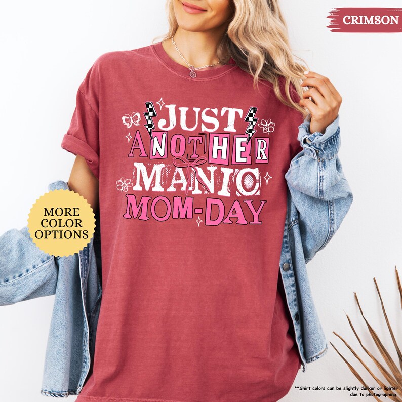 May include: Crimson-colored t-shirt with the text "JUST ANOTHER MANIC MOM-DAY" in a pink and white design. The shirt has short sleeves and a relaxed fit. A "MORE COLOR OPTIONS" button is visible.