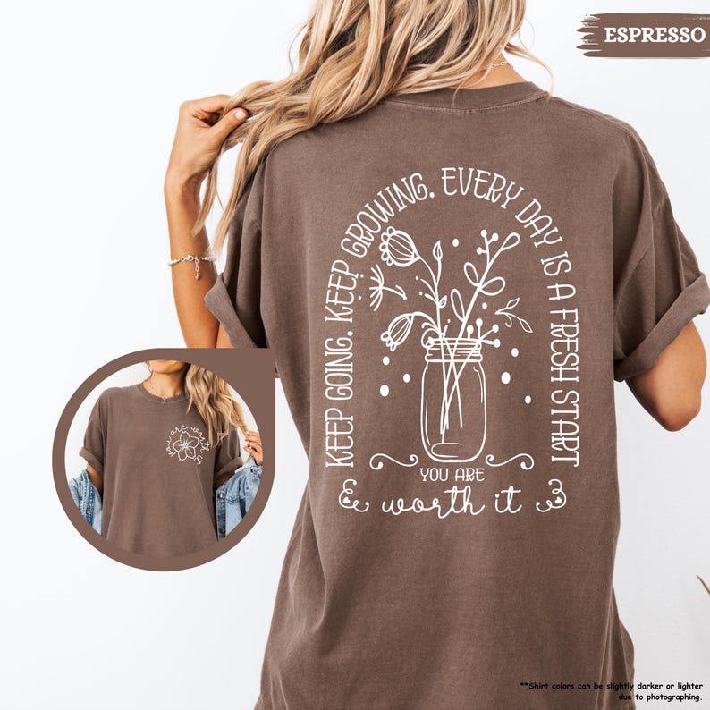 May include: A brown t-shirt with a white graphic design on the back. The graphic features a jar with flowers and the text "Keep going. Keep growing. Every day is a fresh start. You are & worth it."