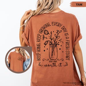 May include: A rust-coloured t-shirt with a black graphic design on the back. The graphic features a mason jar with flowers and the text "Keep going. Keep growing. Every day is a fresh start. You are worth it."