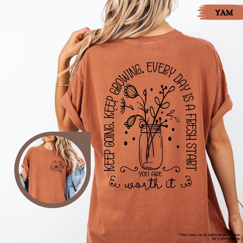 May include: A rust-coloured t-shirt with a black graphic design on the back. The graphic features a mason jar with flowers and the text "Keep going. Keep growing. Every day is a fresh start. You are worth it."