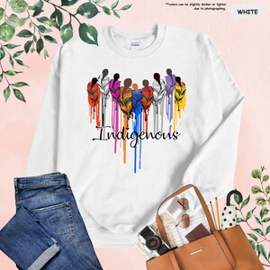 May include: White sweatshirt with a colorful design of stylized figures with feathers and the word "Indigenous" in a dripping paint style.