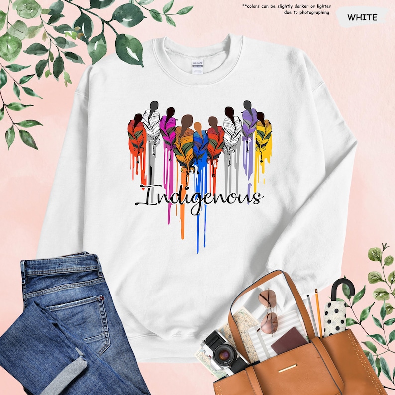 May include: White sweatshirt with a colorful design of stylized figures with feathers and the word "Indigenous" in a dripping paint style.