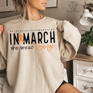 Puede incluir: Una sudadera beige con el texto "#kidneycancerawareness IN MARCH we wear Orange" y una cinta naranja.