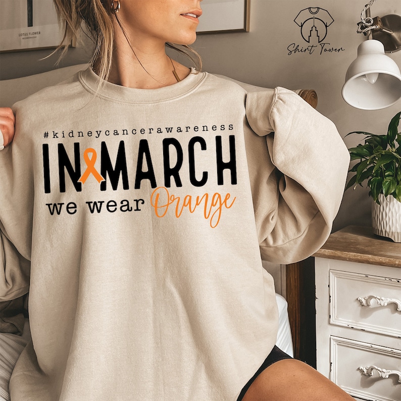 Puede incluir: Una sudadera beige con el texto "#kidneycancerawareness IN MARCH we wear Orange" y una cinta naranja.