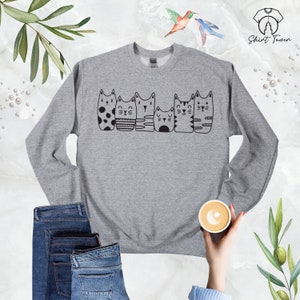 Puede incluir: Sudadera gris con un gr&aacute;fico negro de seis gatos en fila. Los gatos son todos diferentes y tienen caracter&iacute;sticas &uacute;nicas.