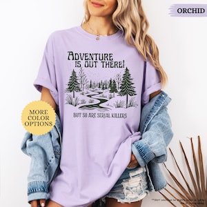 May include: Orchid-colored t-shirt with the text "ADVENTURE IS OUT THERE! BUT SO ARE SERIAL KILLERS" and a graphic of a winding path through a forest. The shirt also has a "MORE COLOR OPTIONS" button.