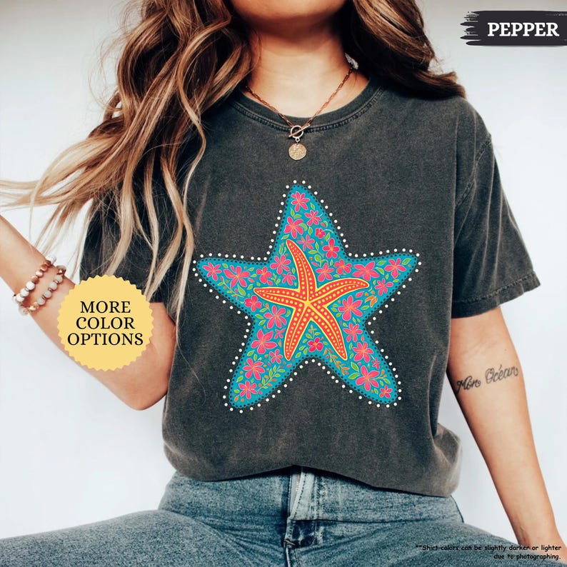May include: A dark gray t-shirt featuring a large, colorful starfish design. The starfish is turquoise with a red and orange starfish in the center, surrounded by pink flowers. The shirt has the word "PEPPER" printed on it and a button that says "MORE COLOR OPTIONS".