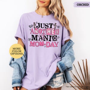 May include: Orchid-colored t-shirt with the text "JUST ANOTHER MANIC MOM-DAY" in a playful font. The design includes butterflies and floral accents. The shirt is a relaxed fit, with rolled-up sleeves.