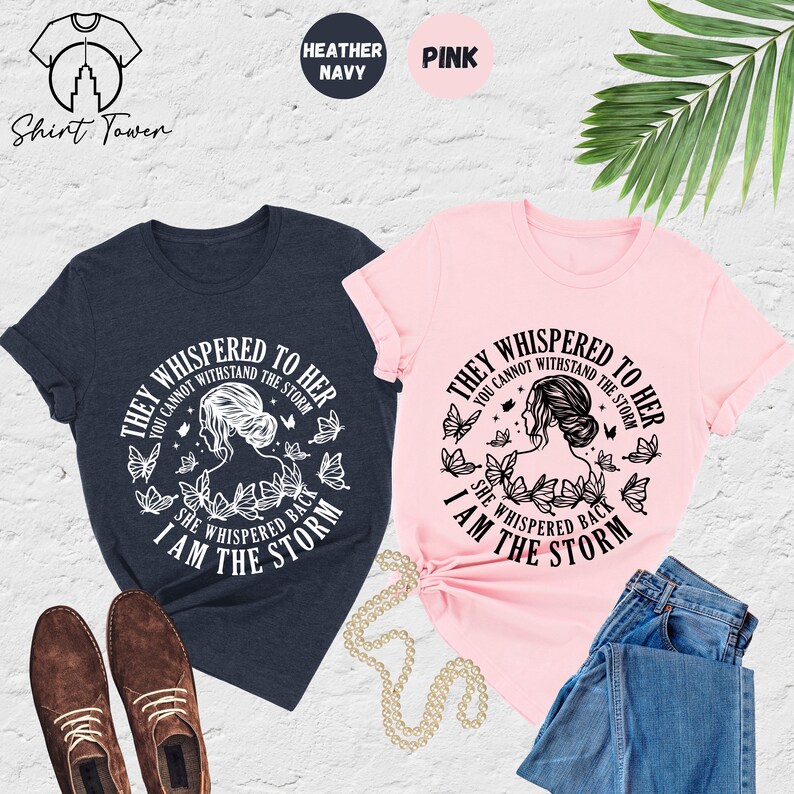 May include: Two t-shirts with a black and white graphic design. The graphic features a woman with butterflies and the text "They whispered to her you cannot withstand the storm she whispered back I am the storm". One t-shirt is heather navy and the other is pink.
