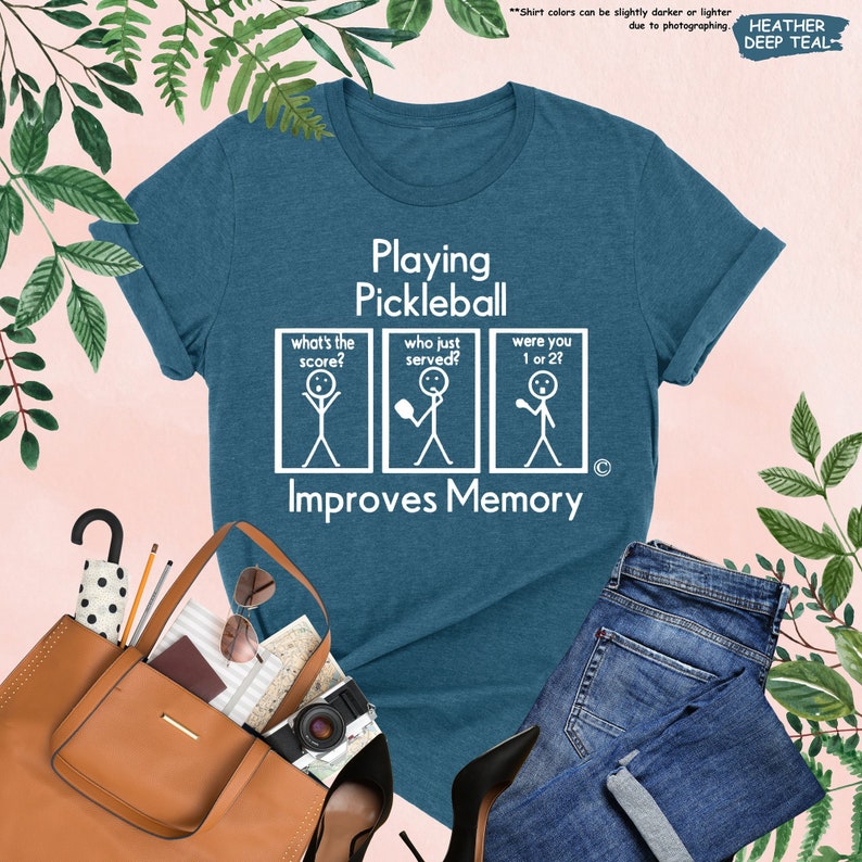 May include: A teal blue t-shirt with white text that reads "Playing Pickleball Improves Memory". The text is accompanied by three stick figure illustrations.