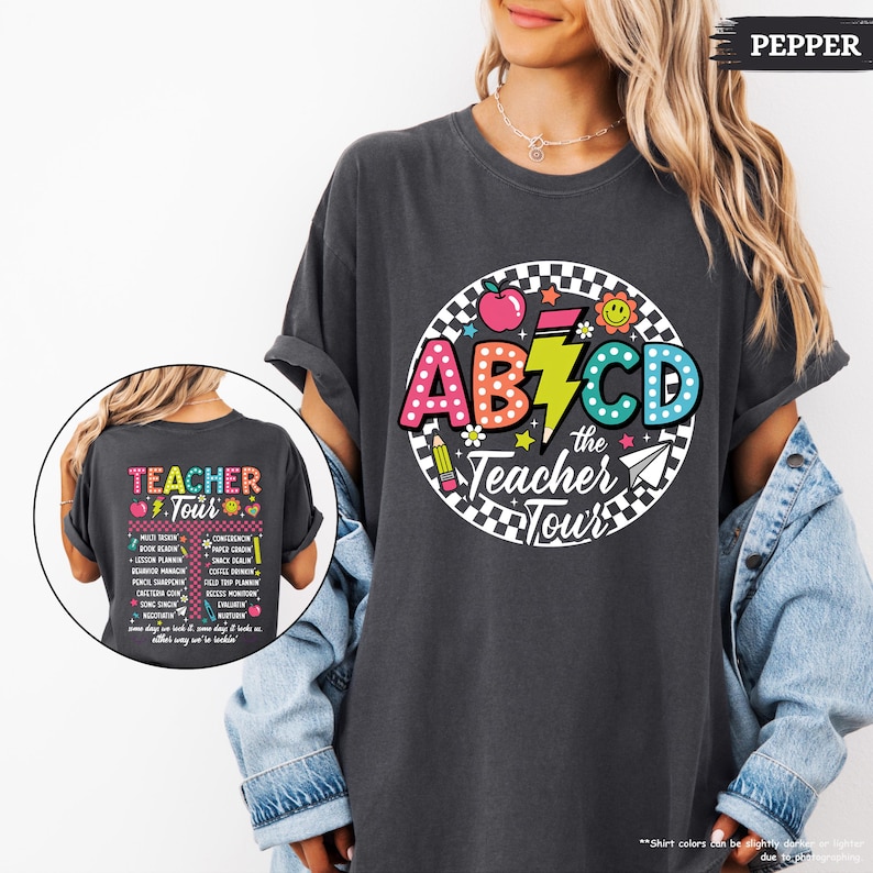 May include: A dark gray t-shirt with a colorful graphic design featuring the letters "ABCD" in a retro style, a lightning bolt, and the text "the Teacher Tour". The back of the shirt has the text "Teacher Tour" with a list of teacher-related tasks.