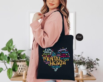 May include: Black tote bag with a colorful graphic design that says "I am okay, I am loving, I am intelligent, I am strong, I am worthy, I am precious, I am a warrior, MENTAL HEALTH, I am loved, I've got this".