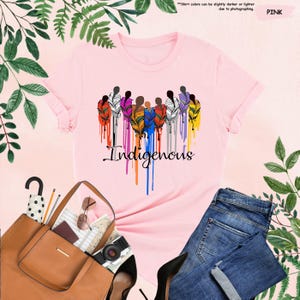 May include: Pink t-shirt with a graphic of a group of people with colorful feathers and the word "Indigenous" printed below.