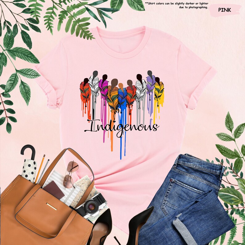 May include: Pink t-shirt with a graphic of a group of people with colorful feathers and the word "Indigenous" printed below.