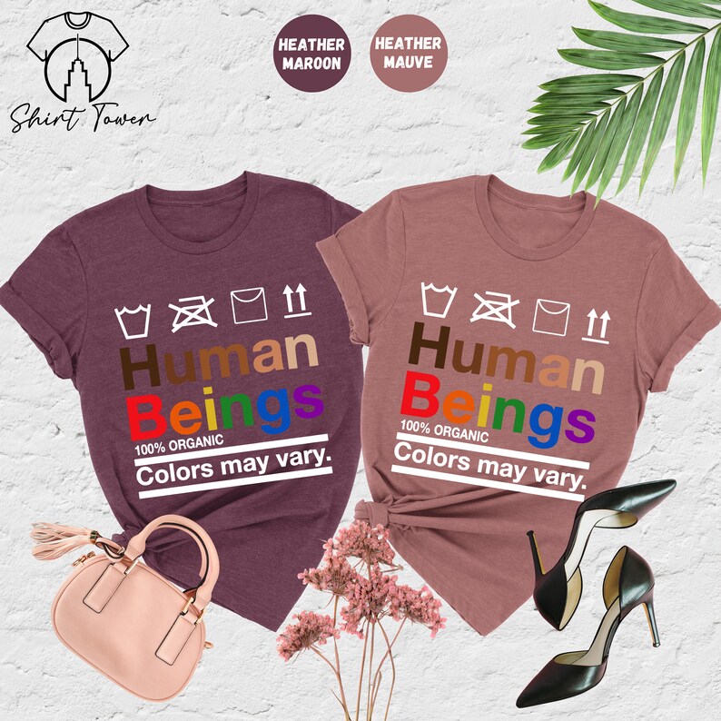 May include: Two t-shirts in heather maroon and heather mauve colors with the text "Human Beings 100% Organic Colors may vary." printed on them. The text is in rainbow colors.