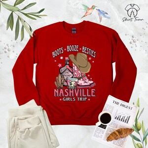 May include: Red sweatshirt with a graphic design of boots, a bottle of whiskey, and a cowboy hat. The text reads "Boots Booze Besties Nashville Girls Trip".