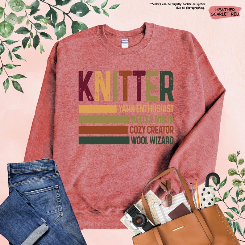 May include: Heather scarlet red sweatshirt with the text "Knitter" in a vintage style font. The text is layered with colorful stripes and the words "Yarn Enthusiast", "Needle Ninja", "Cozy Creator", and "Wool Wizard" are written below.