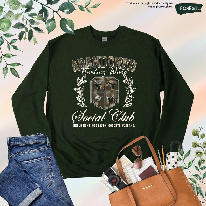 May include: A dark green sweatshirt with the text "Abandoned Hunting Wives Social Club" in white and camouflage lettering. The sweatshirt features a crest with hunting-related symbols and the phrase "Hello Hunting Season. Goodbye Husband."