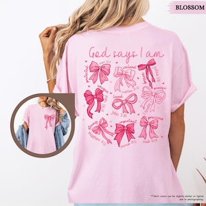 K&ouml;nnte beinhalten: Pinkes T-Shirt mit dem Text "Gott sagt, ich bin" und einem Kreis aus rosafarbenen Schleifen mit Bibelversen. Jede Schleife hat ein Wort wie "gesch&uuml;tzt", "gesch&auml;tzt", "einzigartig", "geliebt", "stark", "wichtig", "kostbar", "m&auml;chtig" und "mutig".