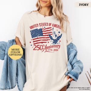 Op de afbeelding: Ivoor kleurig t-shirt met een distressed afbeelding van de Amerikaanse vlag, het Vrijheidsbeeld en een adelaar. De tekst luidt "UNITED STATES OF AMERICA" en "250th Anniversary 1776-2026". Het shirt heeft opgerolde mouwen.