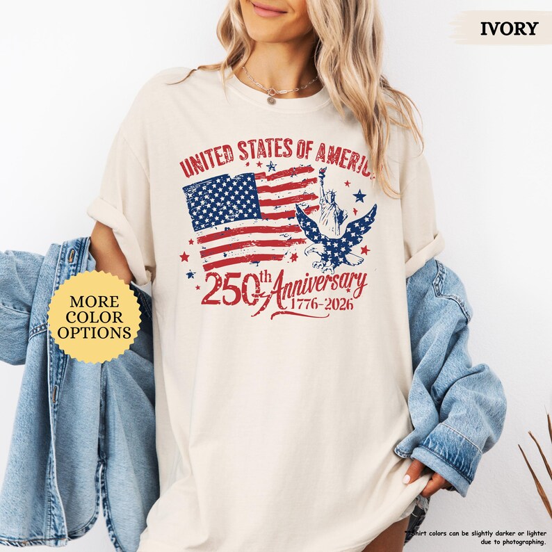 Op de afbeelding: Ivoor kleurig t-shirt met een distressed afbeelding van de Amerikaanse vlag, het Vrijheidsbeeld en een adelaar. De tekst luidt "UNITED STATES OF AMERICA" en "250th Anniversary 1776-2026". Het shirt heeft opgerolde mouwen.