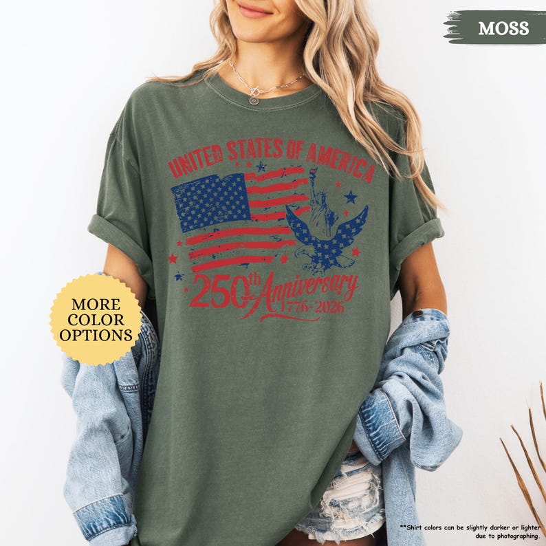 Op de afbeelding: Mosgroen T-shirt met een verweerd Amerikaans vlagontwerp en de tekst "UNITED STATES OF AMERICA" en "250th Anniversary 1776-2026". Het shirt heeft ook een afbeelding van een adelaar en het Vrijheidsbeeld. De mouwen zijn opgerold.