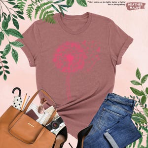 Puede incluir: Una camiseta de color malva con un gran gr&aacute;fico de diente de le&oacute;n rosa. La imagen incluye vaqueros azules, un bolso marr&oacute;n y tacones altos negros. El texto "HEATHER MAUVE" es visible. El color de la camiseta puede variar.