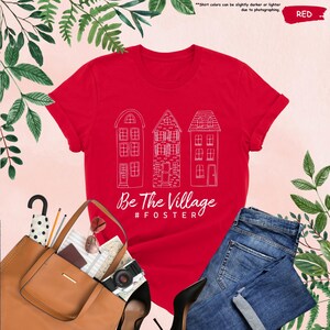 May include: Red t-shirt with a white outline of three houses and the text "Be The Village #FOSTER"