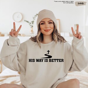 May include: Sand-colored sweatshirt with the text "HIS WAY IS BETTER" in black. The design includes a cross above a curved line. The model is wearing a matching beanie.