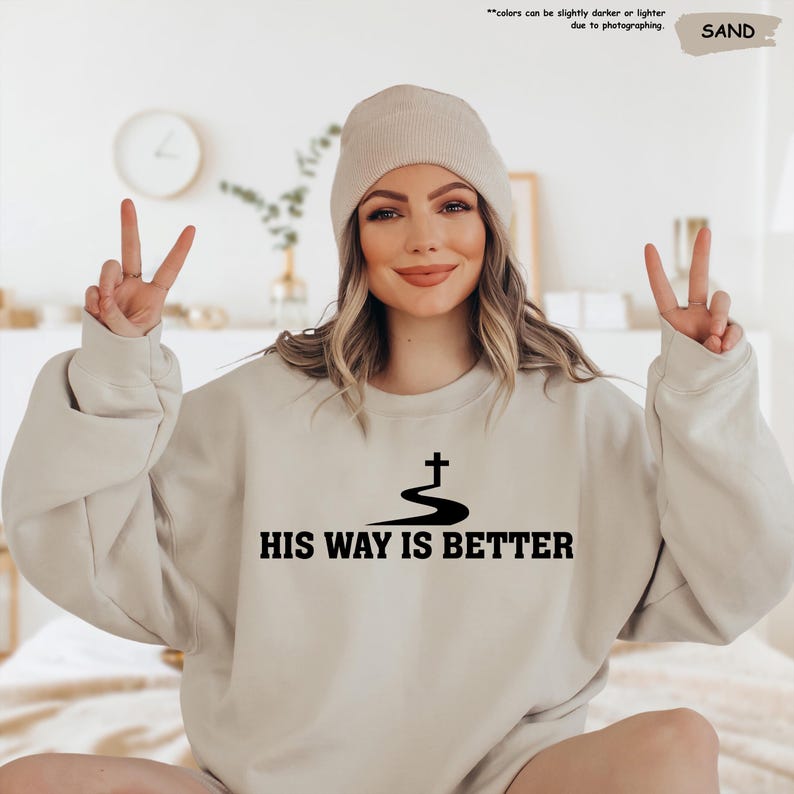 May include: Sand-colored sweatshirt with the text "HIS WAY IS BETTER" in black. The design includes a cross above a curved line. The model is wearing a matching beanie.