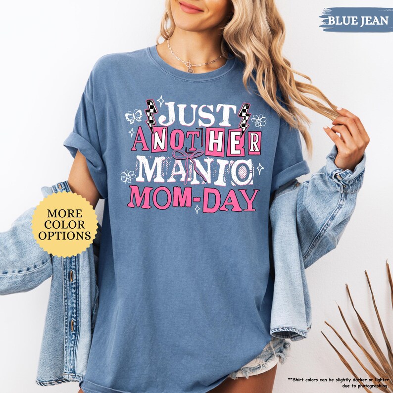 May include: Blue jean-colored t-shirt with the text "JUST ANOTHER MANIC MOM-DAY" in white and pink. The shirt has rolled-up sleeves and a relaxed fit. The words "MORE COLOR OPTIONS" are in a yellow circle.