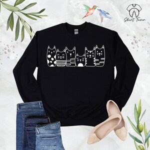 Puede incluir: Sudadera negra con un gr&aacute;fico blanco de cinco gatos en fila, cada uno con un patr&oacute;n diferente. Los gatos miran todos hacia la derecha.