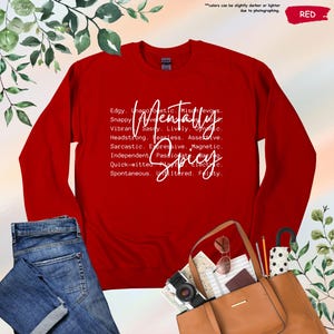 May include: A red sweatshirt with the words "Mentally Spicy" in white script. The design includes a list of adjectives such as "Edgy," "Sassy," and "Fearless." The sweatshirt is paired with blue jeans and a brown tote bag.