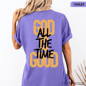 May include: A violet-colored t-shirt with the words "GOD ALL THE TIME GOOD" printed on the back in a combination of yellow and black. The shirt has rolled-up sleeves and a relaxed fit.