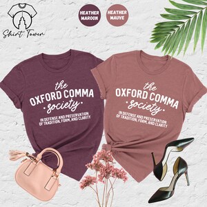 Op de afbeelding: Twee T-shirts in heide-marron en heide-paars met de tekst "the Oxford comma society. In defense and preservation of tradition, form, and clarity" in wit gedrukt.