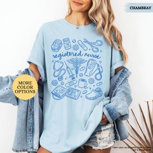 May include: Light blue t-shirt with a blue graphic of medical tools and the words "registered nurse." The design includes a stethoscope, syringe, and other medical equipment. The shirt has short sleeves and a crew neck.