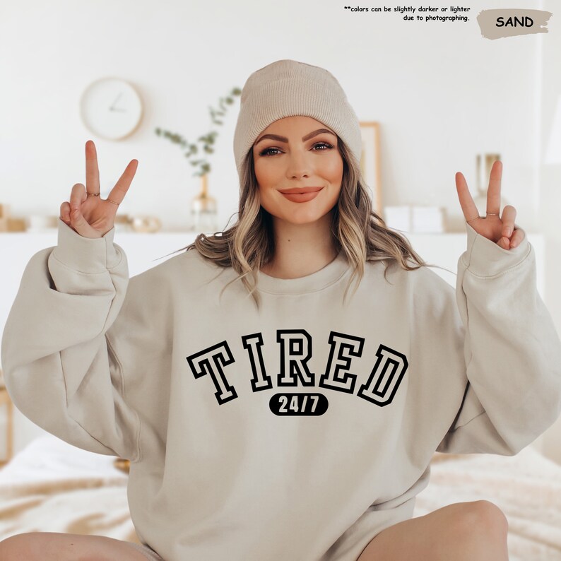 a woman sitting on a bed wearing a sweatshirt that says tired