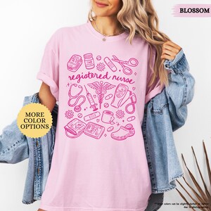 May include: Light pink t-shirt with a pink graphic of medical tools and the words "registered nurse." The design includes a stethoscope, syringe, and other medical equipment. The shirt also has the text "MORE COLOR OPTIONS" and "BLOSSOM."