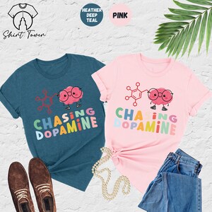 Pu&ograve; includere: Due magliette con un cervello di cartone animato che indossa occhiali e tiene una molecola. Il testo sulle magliette recita "Chasing Dopamine" in colori arcobaleno. Una maglietta &egrave; blu scuro melange e l'altra &egrave; rosa.
