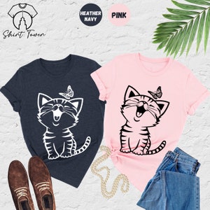 May include: Two t-shirts with a graphic of a smiling cat with a butterfly above it. The t-shirts are in heather navy and pink.
