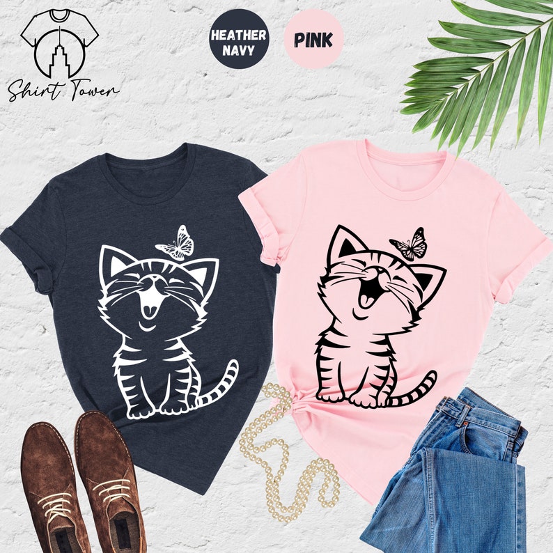 May include: Two t-shirts with a graphic of a smiling cat with a butterfly above it. The t-shirts are in heather navy and pink.