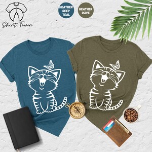 May include: Two t-shirts with a white graphic of a smiling cat with a butterfly above it. The t-shirt on the left is heather deep teal and the t-shirt on the right is heather olive.