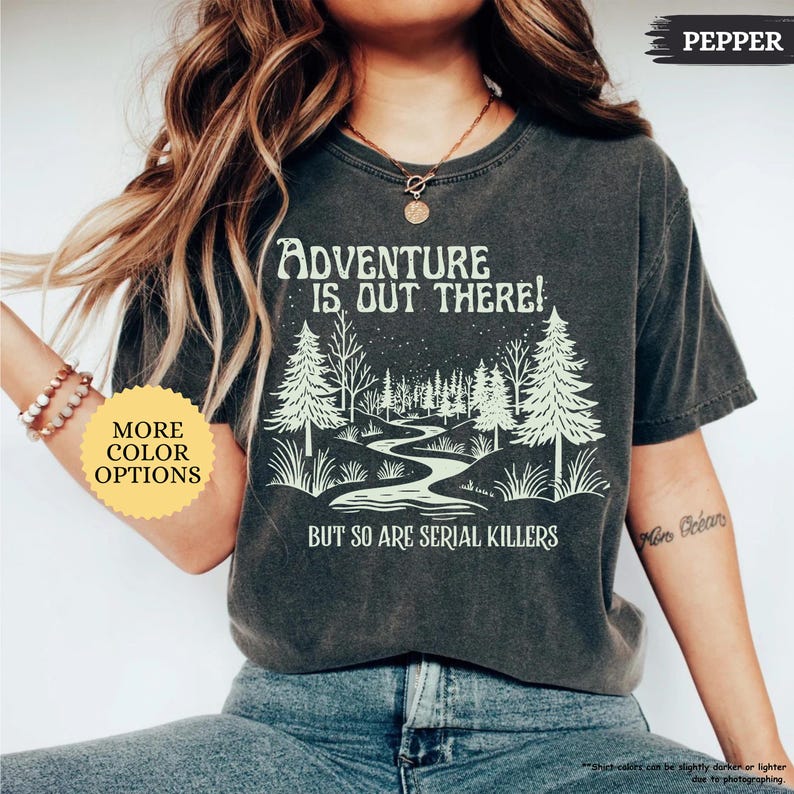 May include: Dark gray t-shirt with the text "ADVENTURE IS OUT THERE! BUT SO ARE SERIAL KILLERS" in white. The graphic features a forest scene with a winding river. The shirt has a vintage, worn look.
