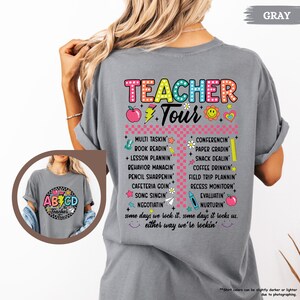 May include: Gray t-shirt with a colorful graphic that says "Teacher Tour" and lists tasks teachers do, such as "Multi Tasking", "Book Readin", "Lesson Plannin", "Behavior Managnin", "Pencil Sharpenin", "Cafeteria Goin", "Song Singin", "Negotiatin", "Conferencing", "Paper Gradin", "Snack Dealn", "Coffee Drinkin", "Field Trip Plannin", "Recess Monitorin", "Evaluatin", and "Nurturin". The graphic also includes a red apple, a yellow sun, and a pink heart.