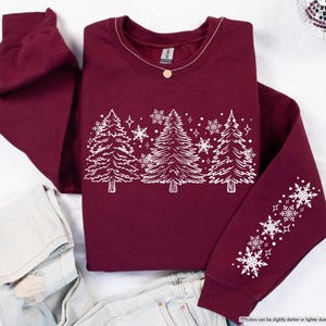 May include: A maroon sweatshirt with a white design of stylized Christmas trees and snowflakes. The design is on the front of the sweatshirt and the sleeves have a line of snowflakes.