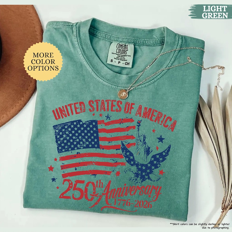 Op de afbeelding: Lichtgroen T-shirt met een patriottisch ontwerp. Het shirt heeft de tekst "UNITED STATES OF AMERICA" boven een afbeelding van de Amerikaanse vlag, het Vrijheidsbeeld en een adelaar. De tekst "250th Anniversary 1776-2026" staat ook op het shirt.